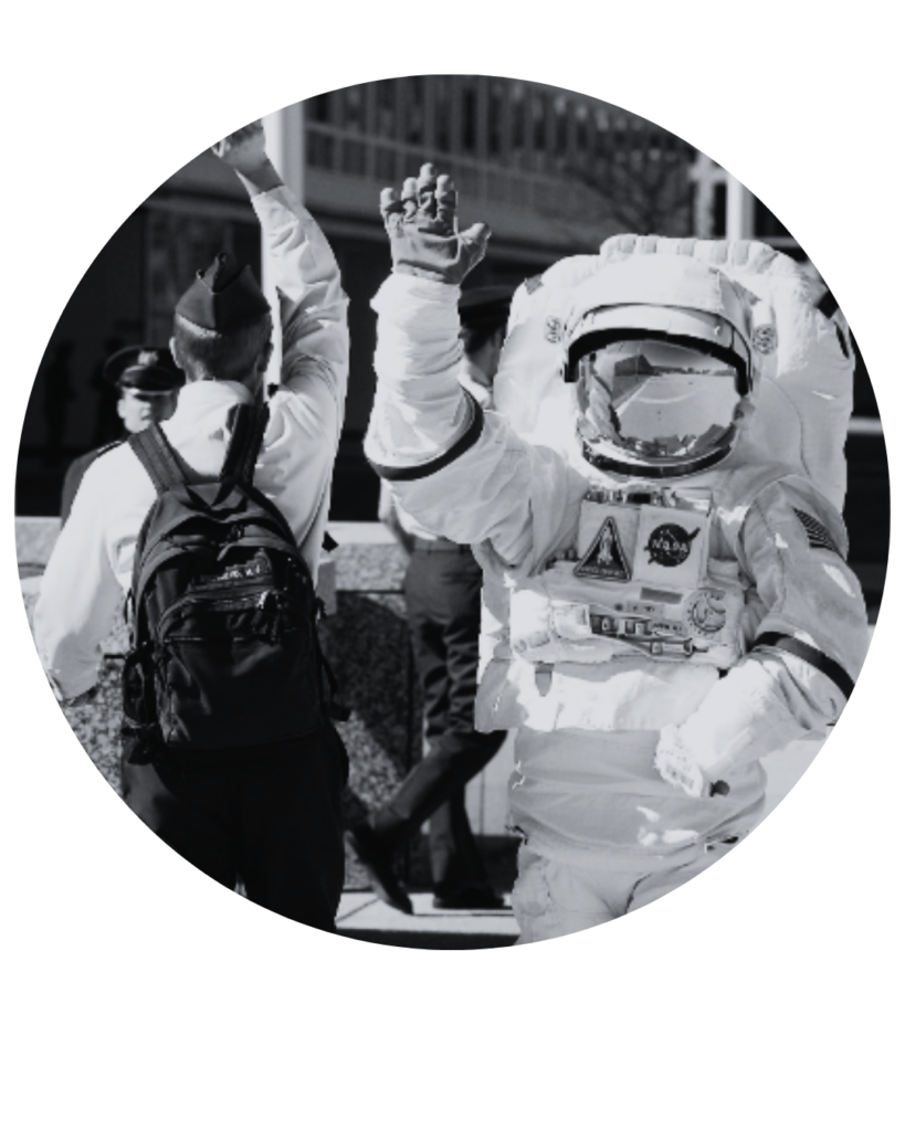Astronauta com traje completo da NASA acenando em uma rua urbana, ao lado de uma pessoa de costas com mochila.