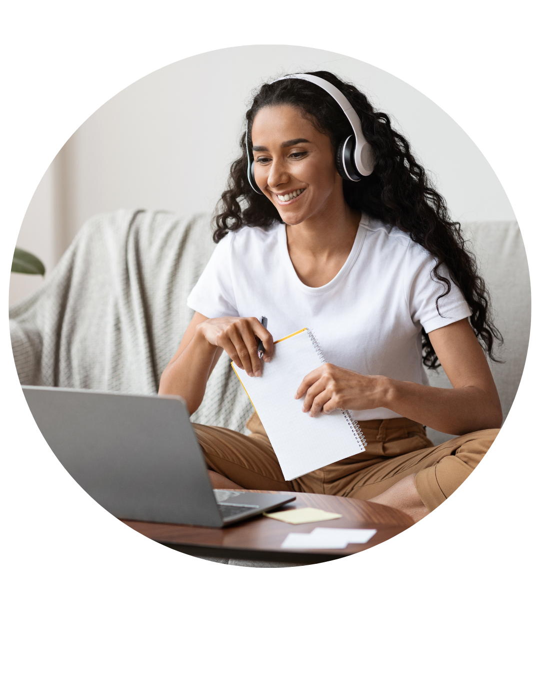 Mulher sorridente usando fones de ouvido e escrevendo em um caderno enquanto olha para a tela do laptop.